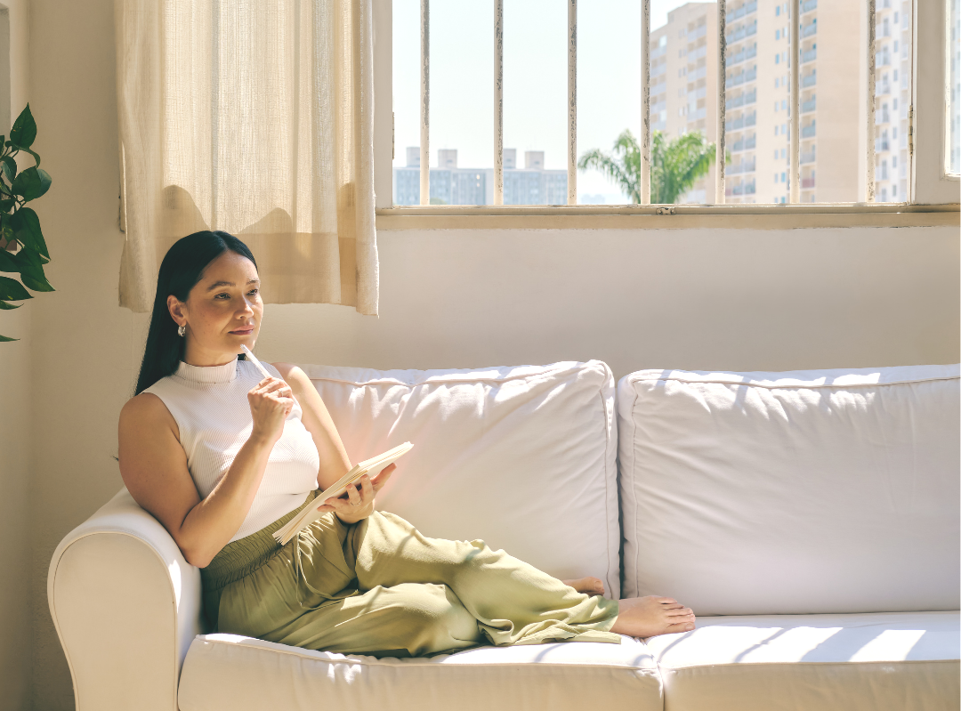 A woman sitting on a white couch in a sunlit room, reading a book and holding a pen, with a window showing city buildings outside.