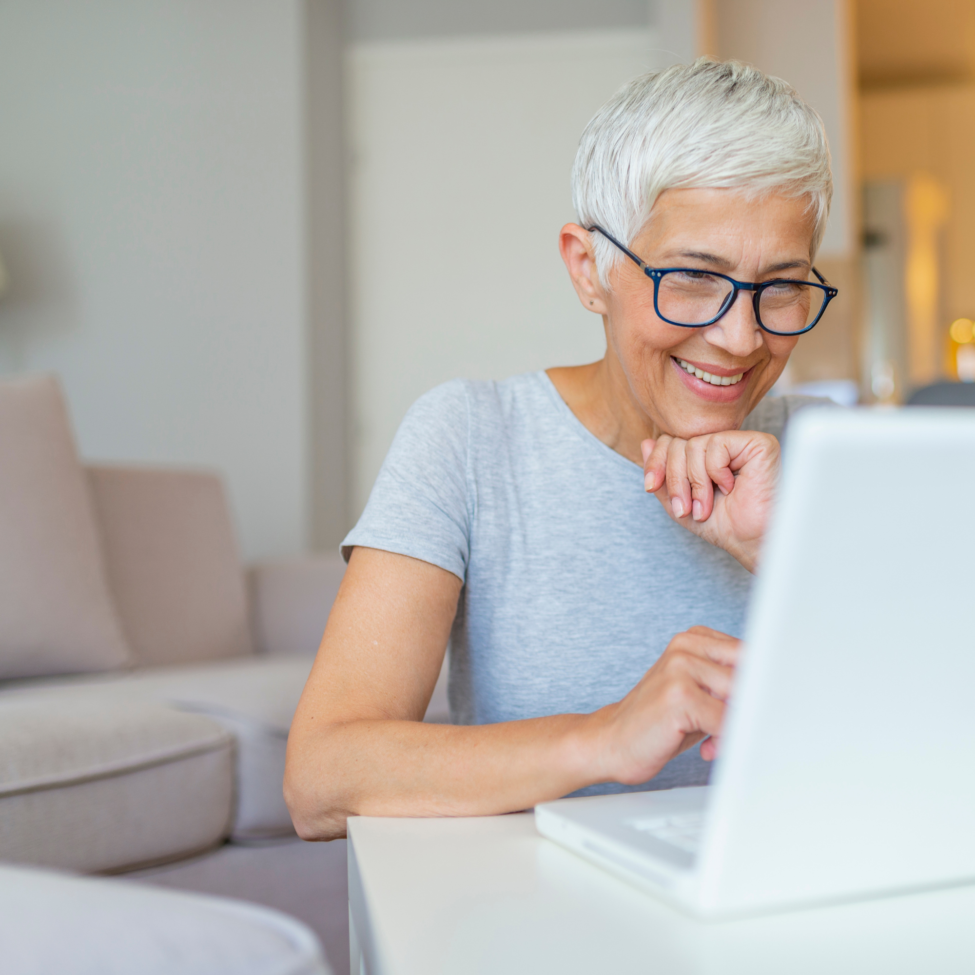 A smiling older woman with short gray hair and glasses, sitting at a desk with a computer, in a bright indoor setting.