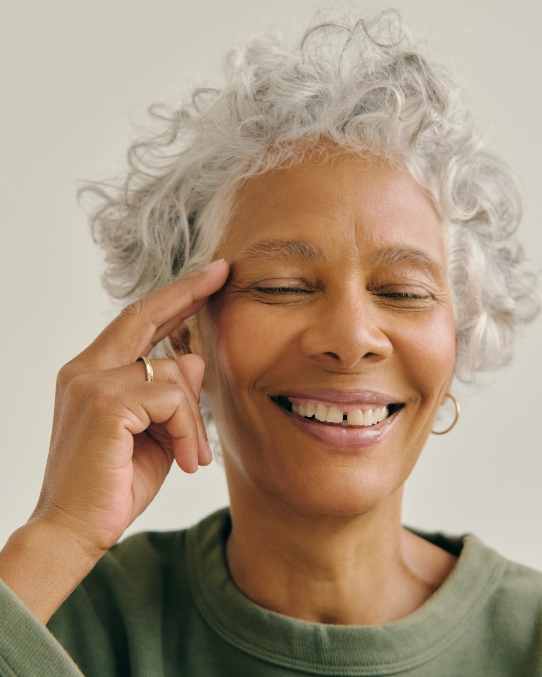 A woman with short, curly gray hair smiling with eyes closed, touching her forehead feeling well.