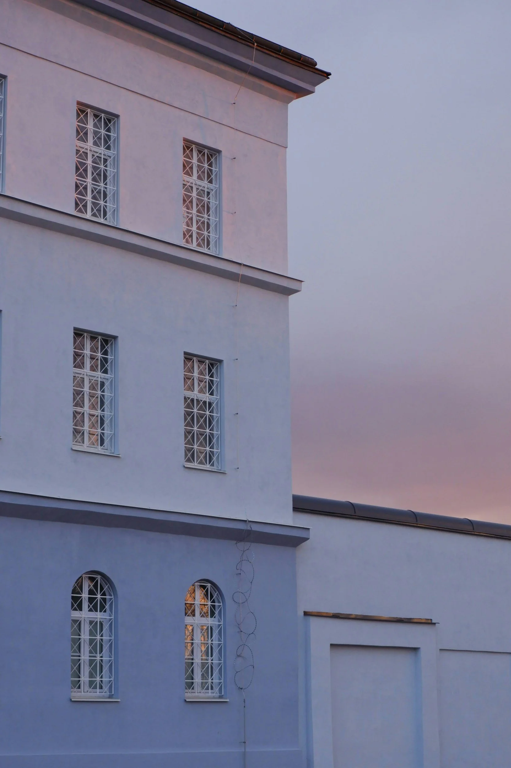 Side of a white building with multiple windows with decorative metal bars, and a sky with pink and purple hues expected at sunset.