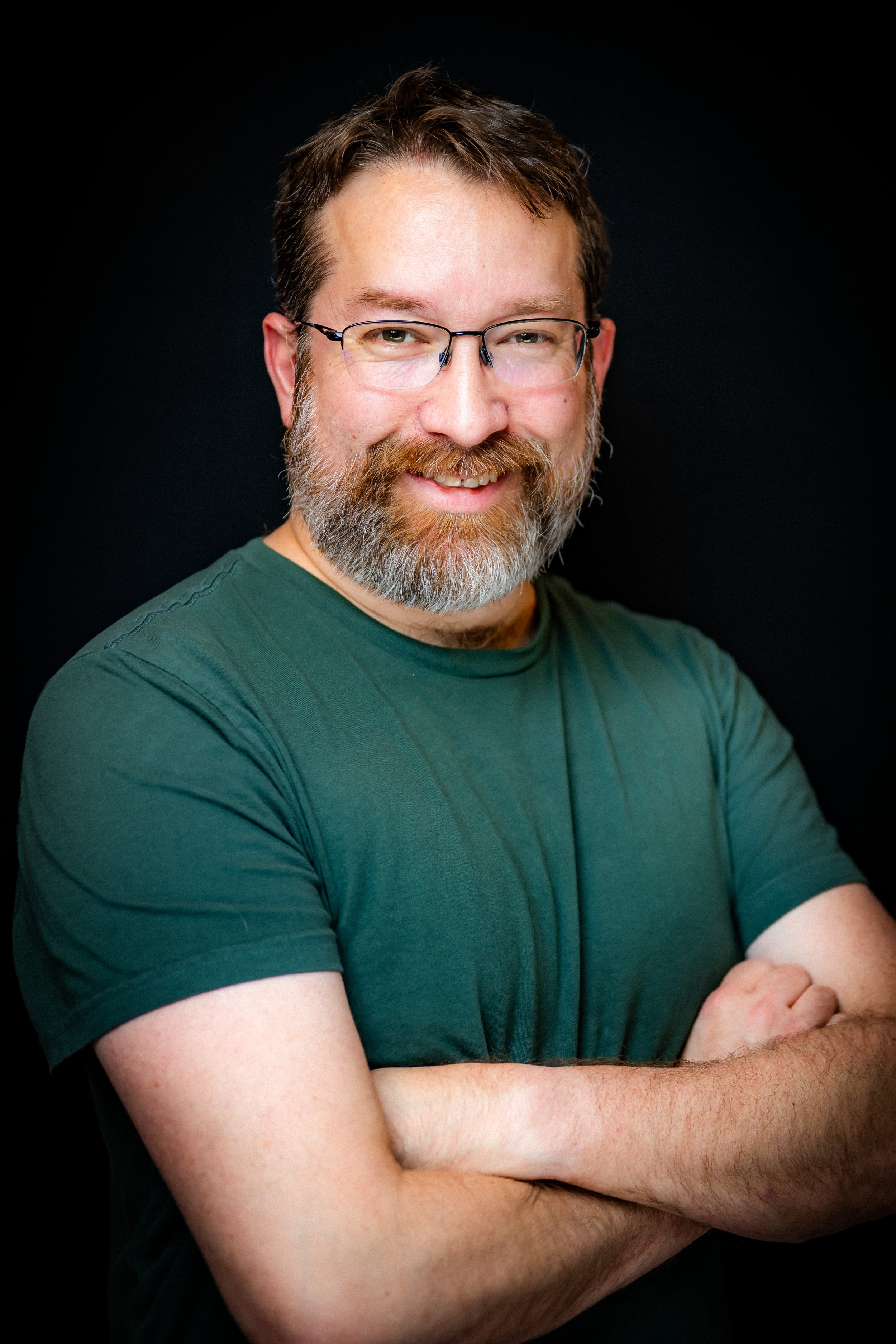 Portrait of a middle-aged man with glasses, beard, and mustache, smiling with arms crossed, wearing a green T-shirt against a black background.