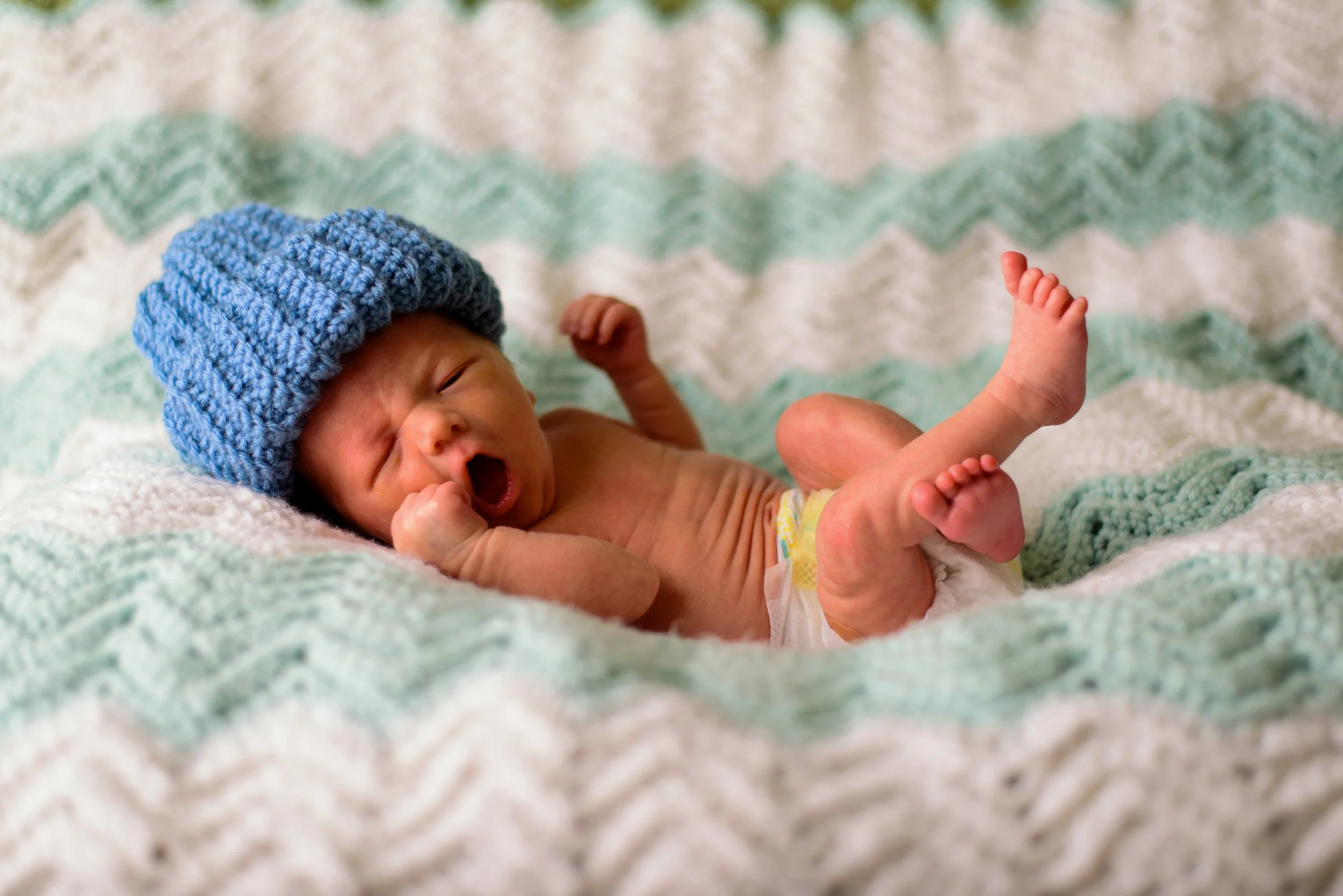 Newborn baby lying on a knitted blanket, wearing a blue knit hat, yawning with eyes closed, one hand near their face and legs raised.
