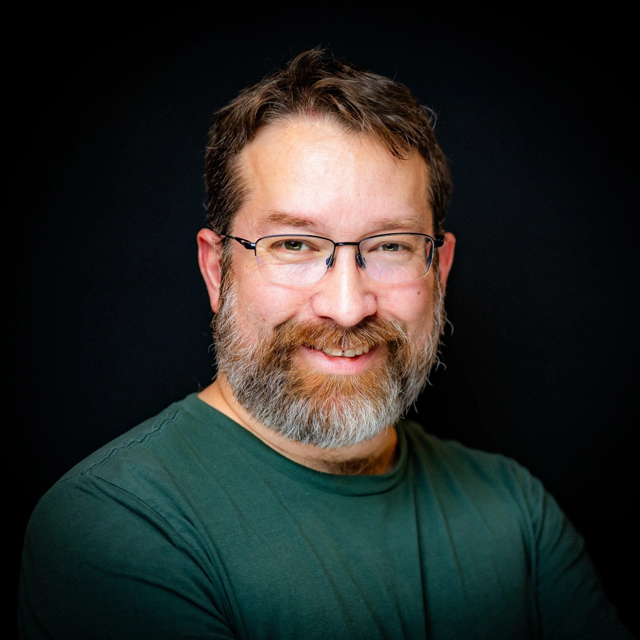 Portrait of a smiling man with glasses, a beard, and mustache, wearing a green shirt, against a dark background.