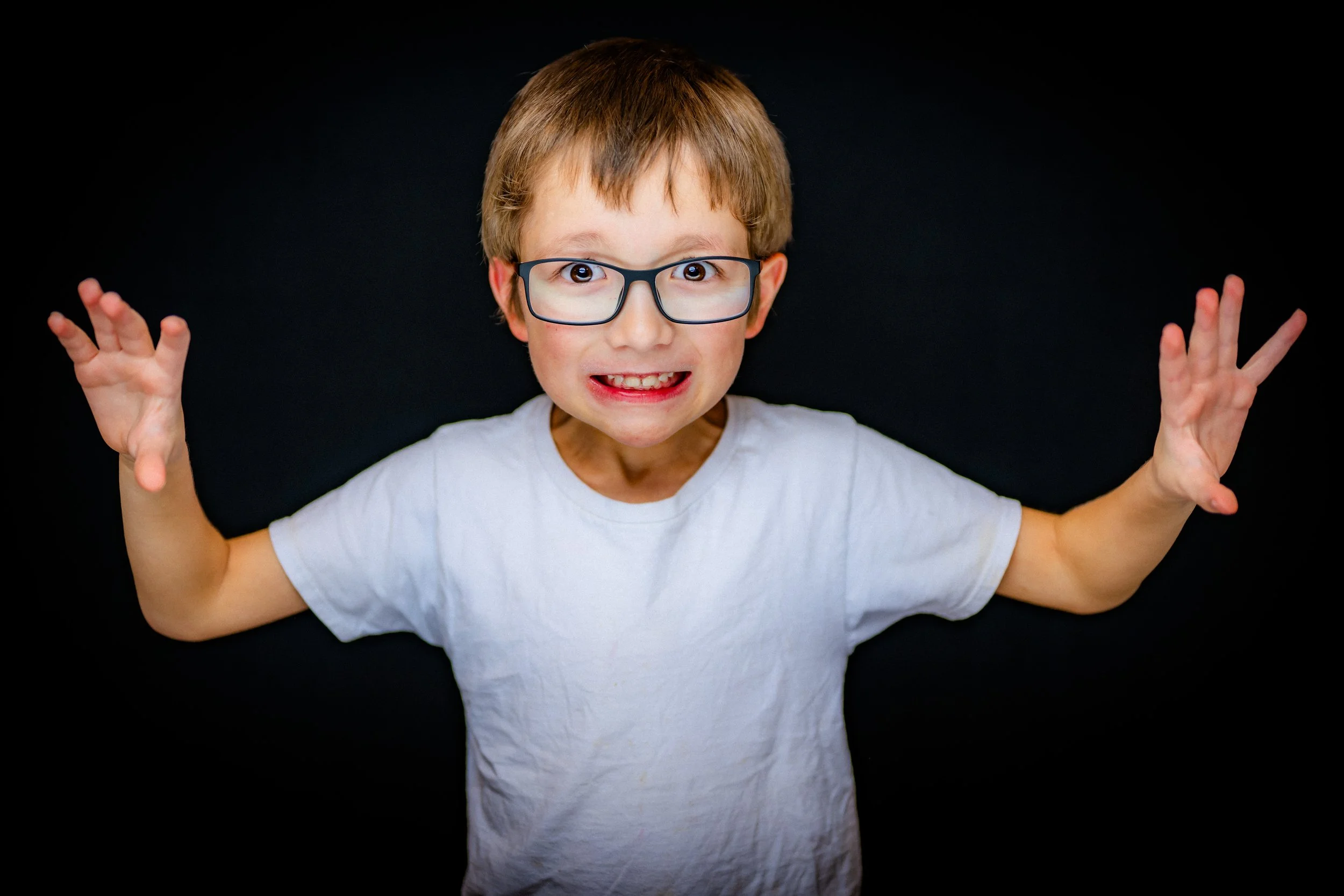 Young boy with glasses, smiling, arms raised, wearing a white t-shirt against a black background.