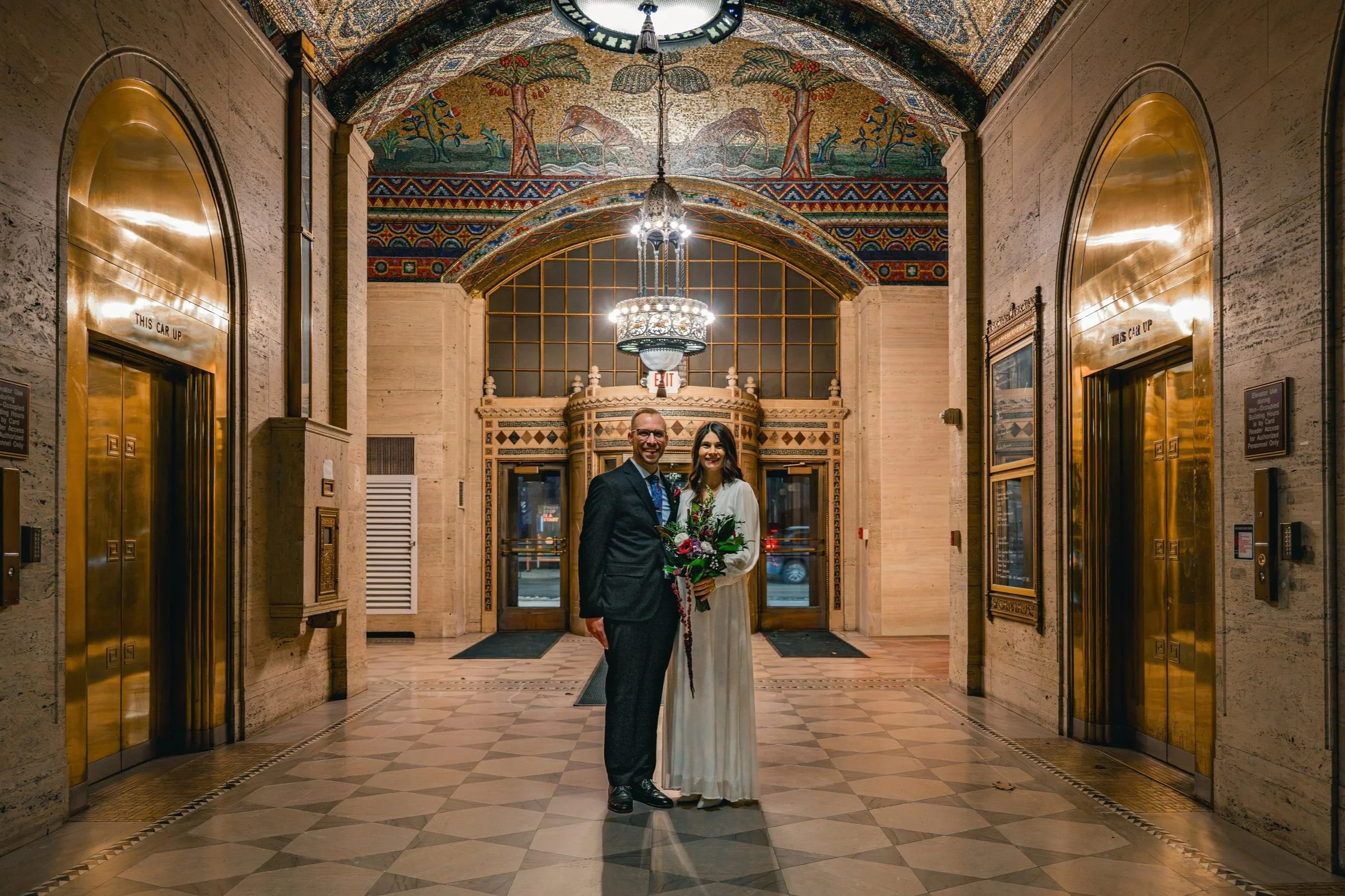 A man and woman standing in front of an ornate lobby, smiling at the camera. The woman is holding a bouquet of flowers. The lobby has a decorative tiled ceiling, chandeliers, and two elevator doors with brass finish.