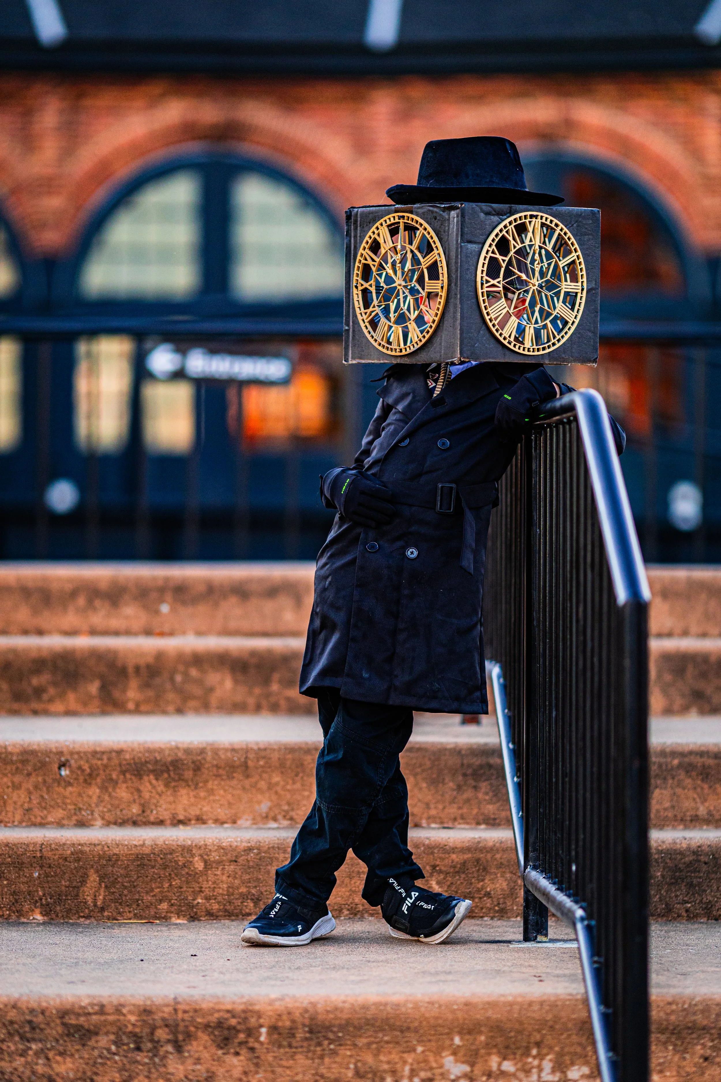 A child dressed in black leaning against railing on steps, wearing a box-shaped costume with clock faces on the sides and a black hat.