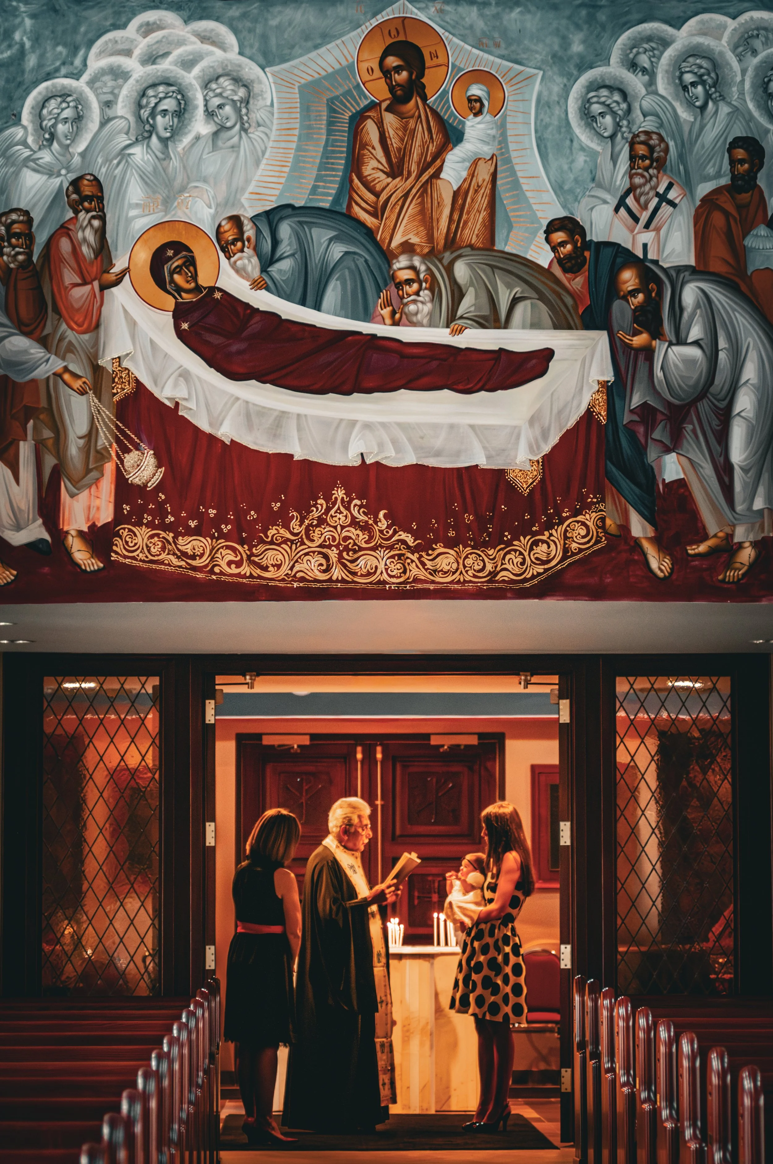 An ornate religious mural depicting the death of Jesus Christ above a church altar, with women and men surrounding His body, and a scene of a baptism or religious ceremony taking place inside the church below, with three women, a priest, and an infant involved in the ceremony.