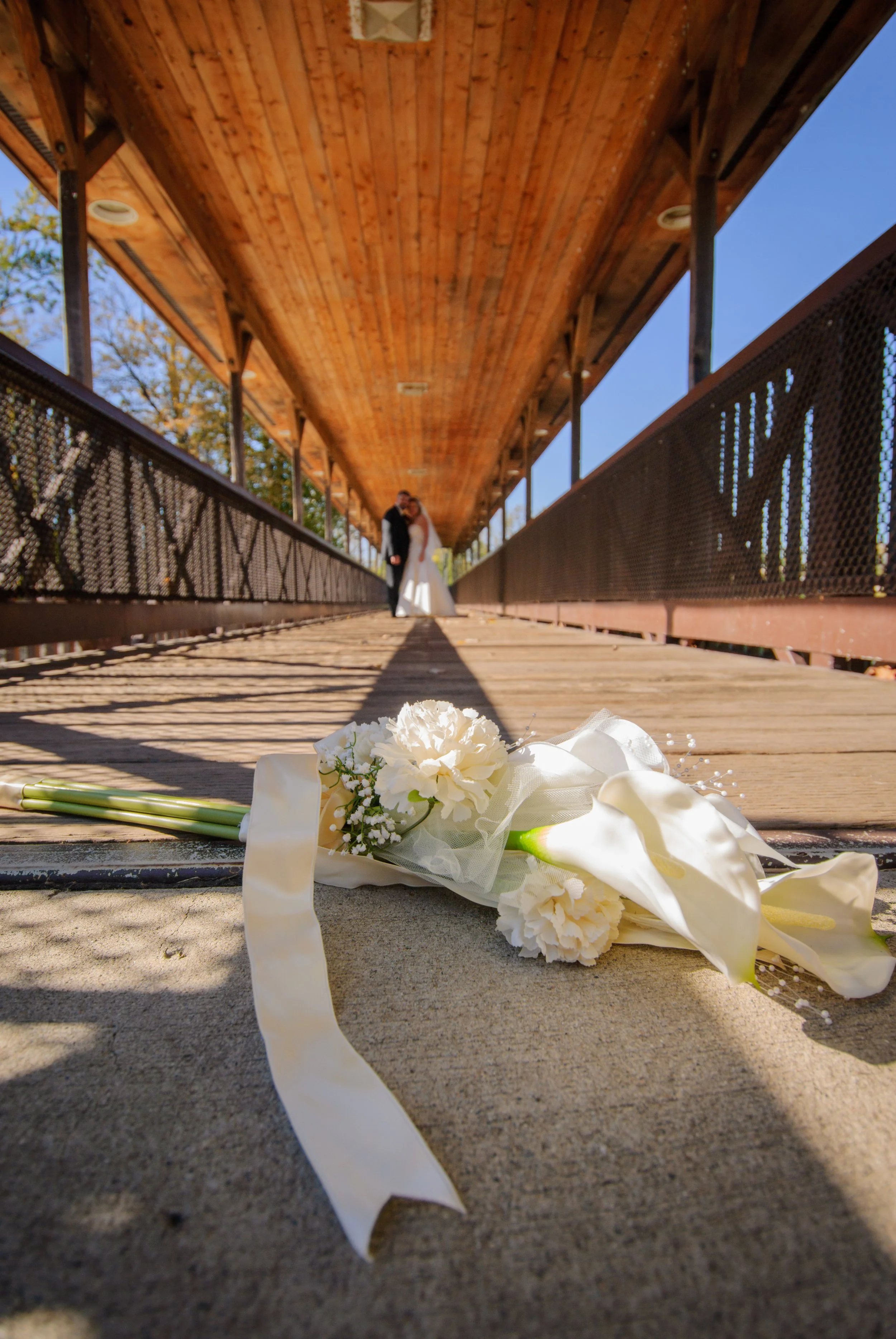 Wedding bouquet with white roses and calla lilies lying on a wooden bridge, with a bride and groom kissing in the background under a covered wooden structure.