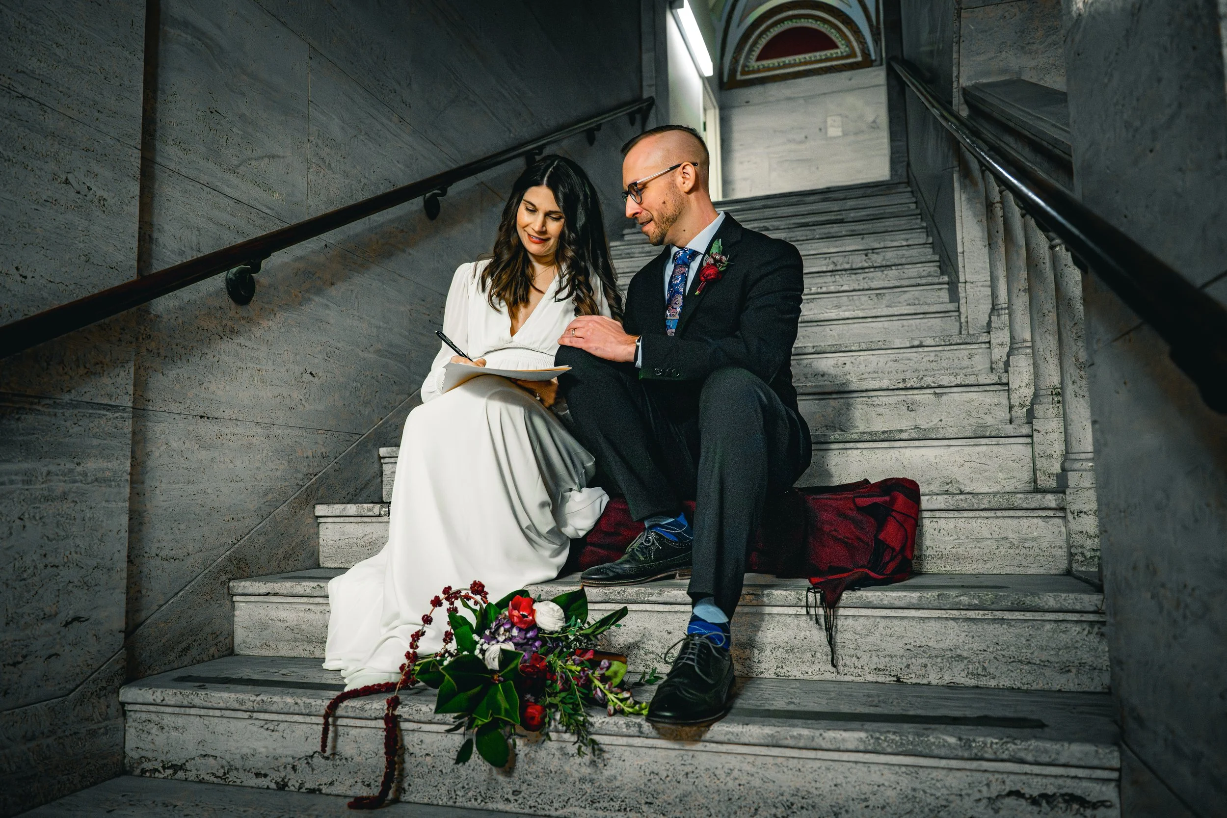 A man and woman sitting on a staircase, the woman wearing a white dress and the man dressed in a dark suit with a colorful tie, both examining a notebook; a bouquet of flowers is on the steps in front of them.