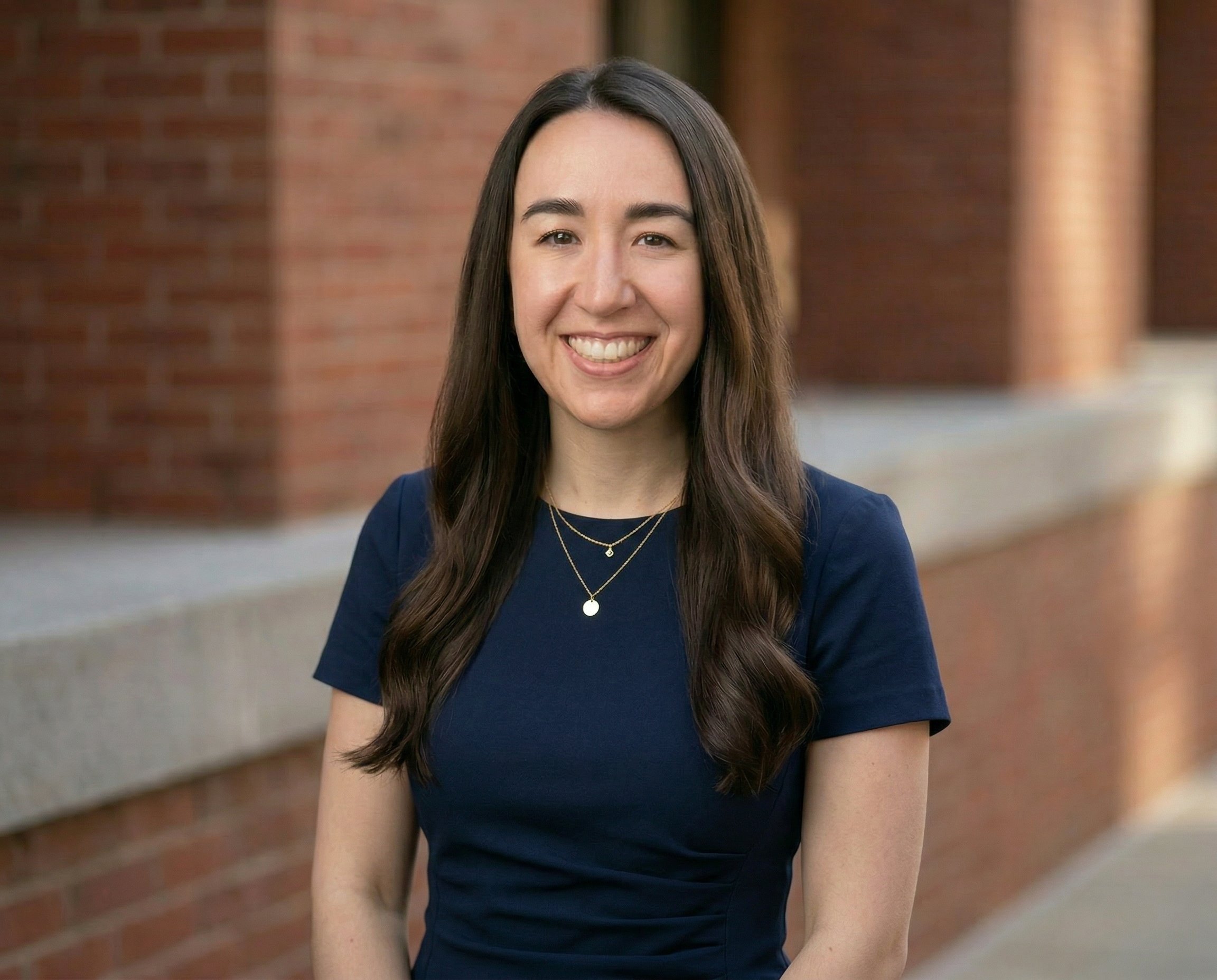 Young woman with long brown hair smiling outdoors in front of a brick building, wearing a navy blue dress and layered necklaces.