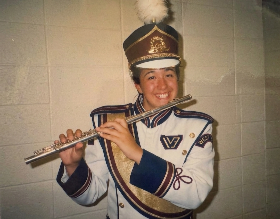 A young woman in a marching band uniform holding a flute and smiling in front of a beige wall.