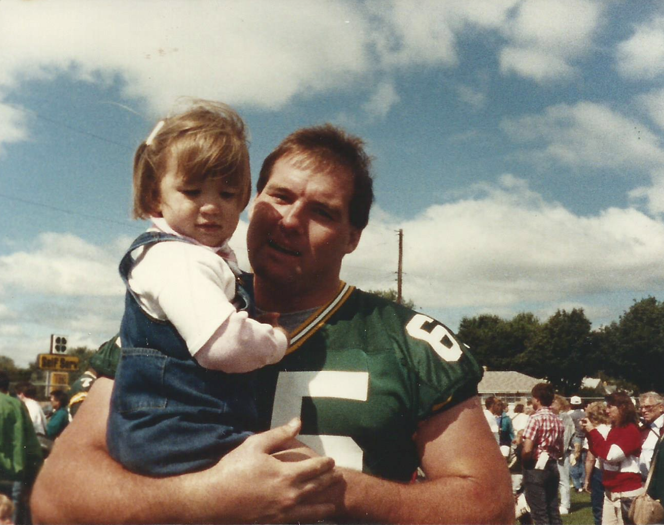 A man in a green sports jersey holding a young girl in a blue dress outside on a cloudy day, with a crowd of people in the background.