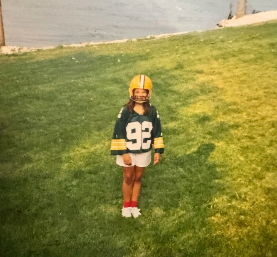 A young girl standing on a grassy hill in a Green Bay Packers football uniform, wearing a helmet and shorts.