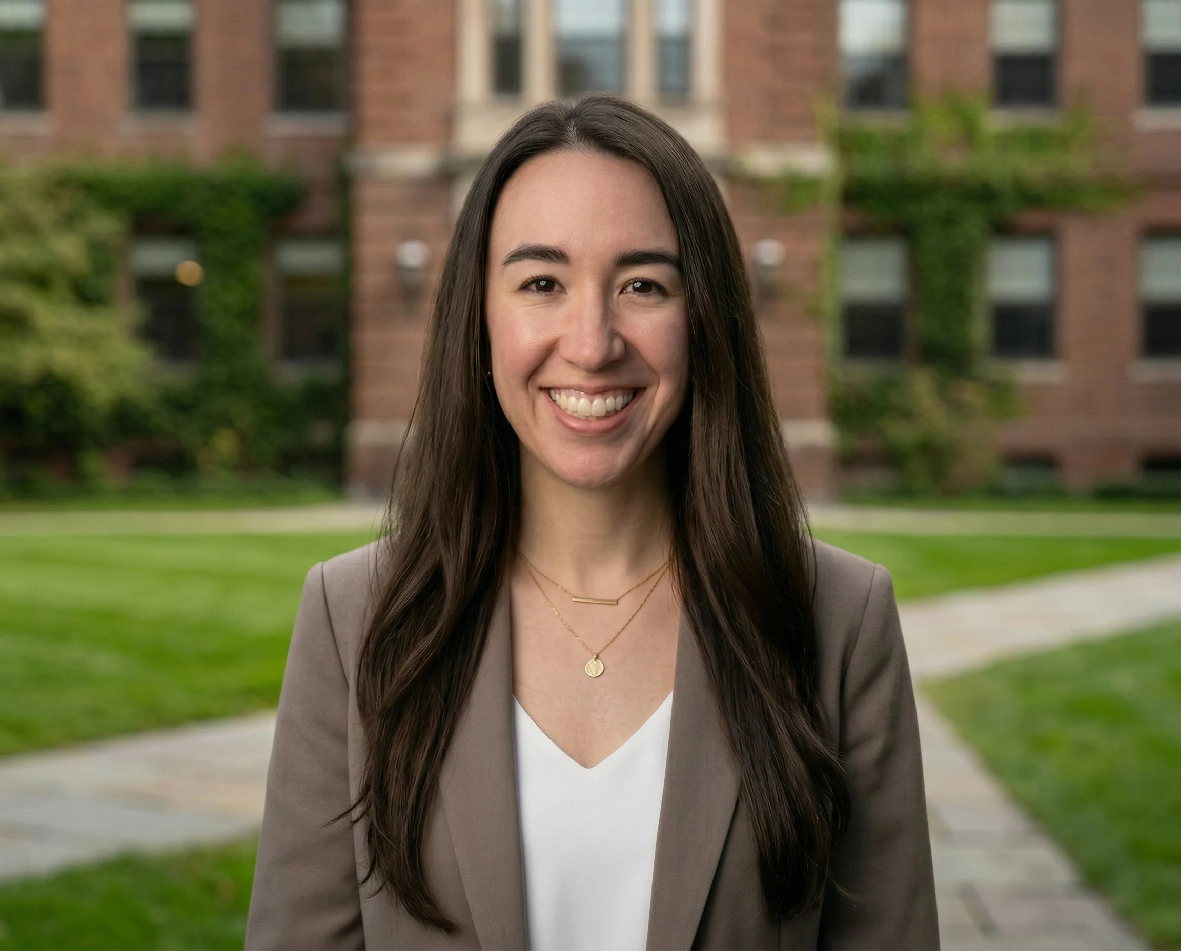 A woman with long dark hair smiling outdoors in front of a brick building and greenery.