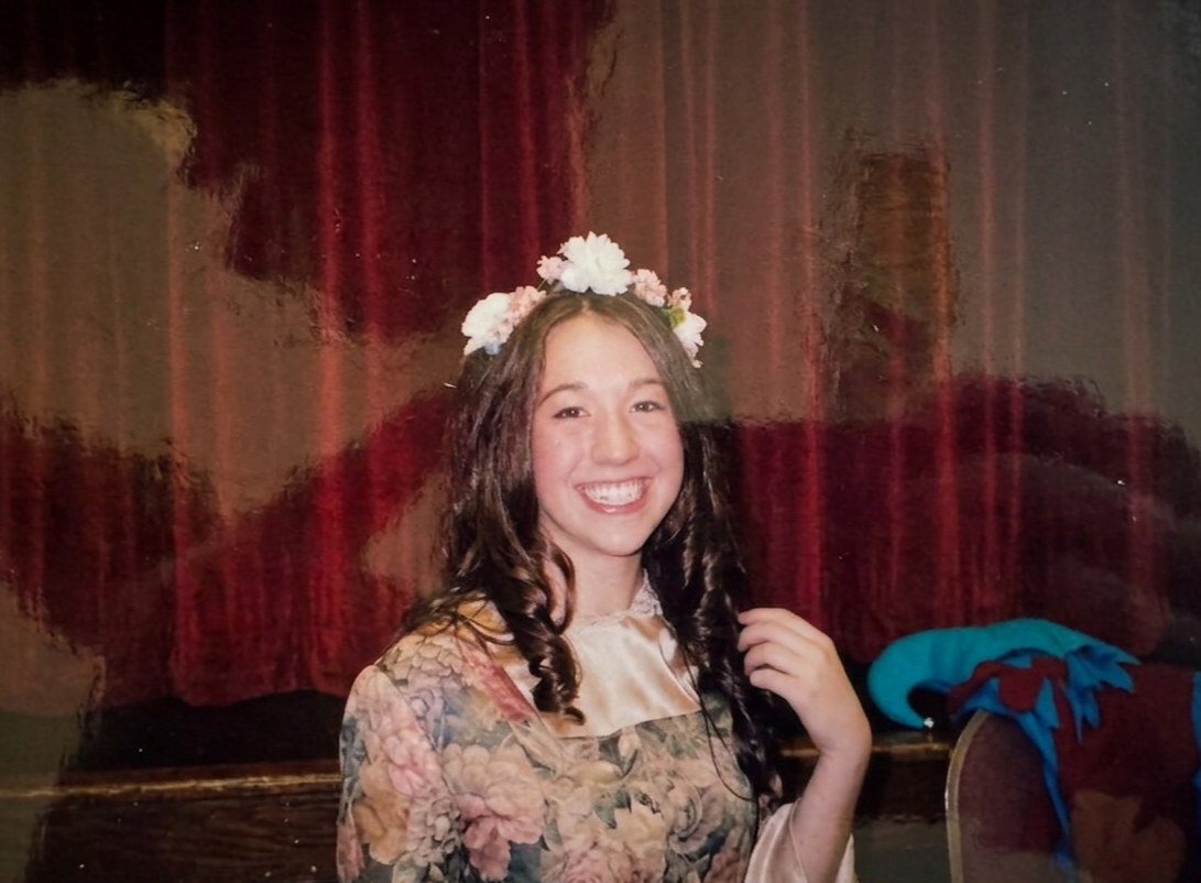 A young woman with long dark hair wearing a floral dress and a flower crown, smiling.