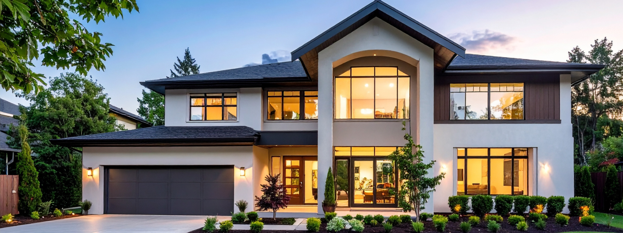 Modern two-story house with large windows, a gray garage door, landscaped front yard with small trees and bushes, and warm interior lighting visible through the windows, during dusk.