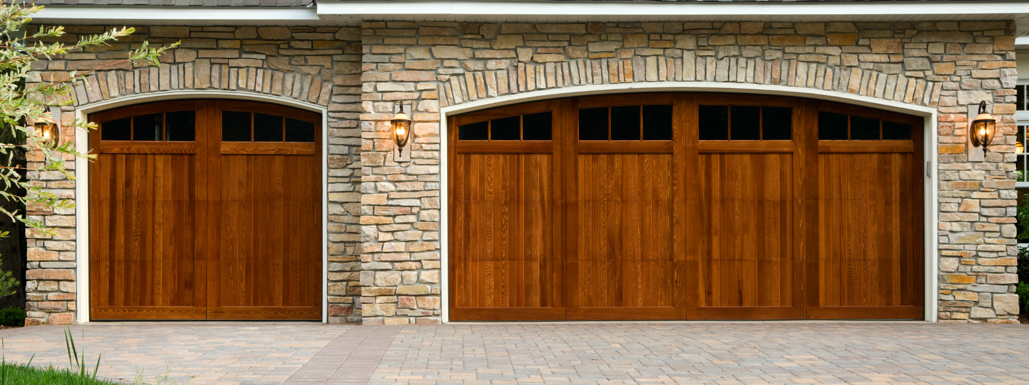 Double garage doors made of wood with stone facade and outdoor wall-mounted lanterns on each side.