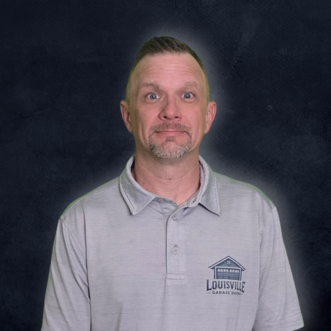 A man with short hair, a goatee, and a surprised expression, wearing a light gray polo shirt with a Louisville Garage Doors logo, standing against a dark textured background.