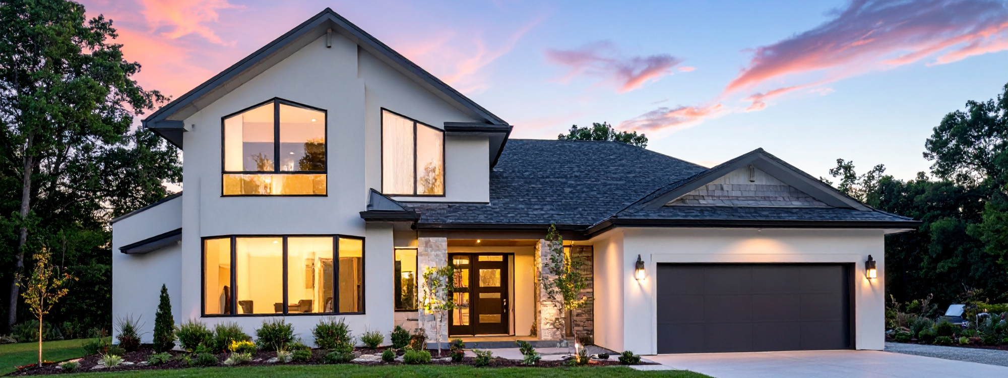 Modern two-story house with large windows, a dark garage door, and exterior lighting, surrounded by trees and landscaped lawn at sunset.