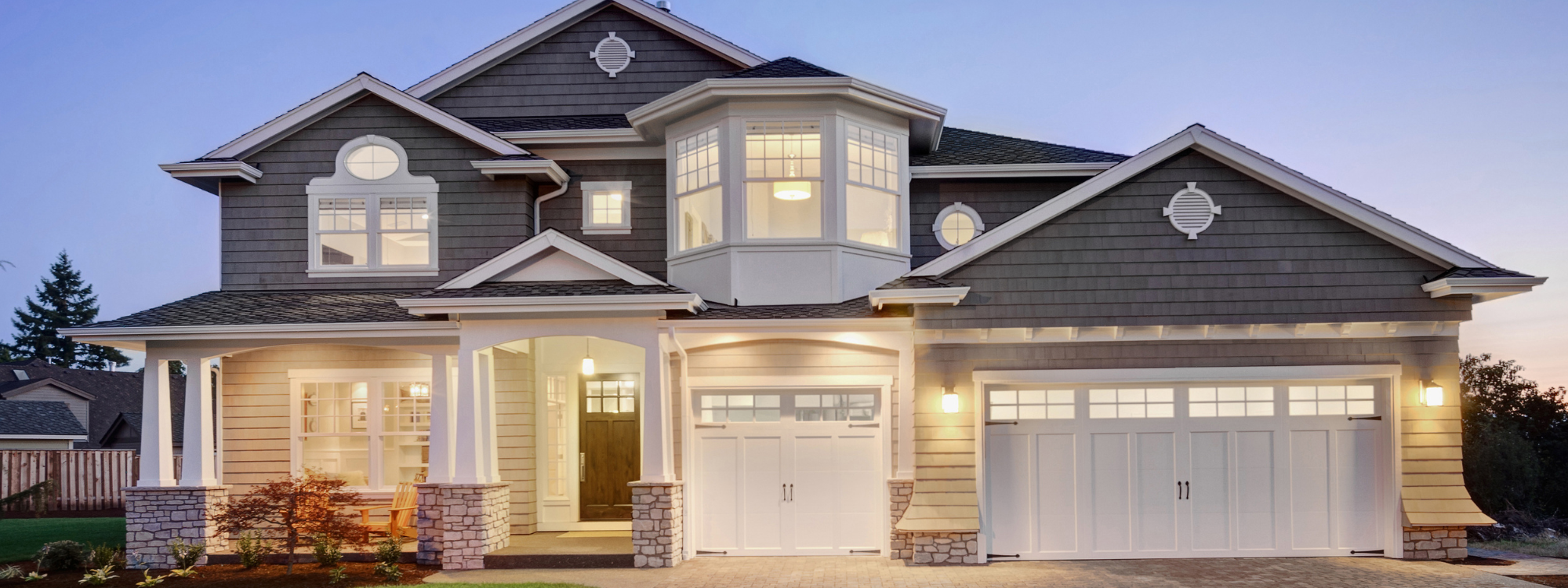 A large two-story house with a gray exterior, white trim, and a three-car garage, illuminated with exterior lights during dusk.