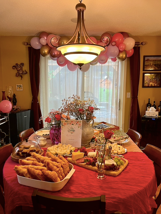 A decorated dining room with a table set for a celebration, featuring a centerpiece of flowers, cheese, fruit, and pastries. Pink, gold, and purple balloons hang from a chandelier above the table, and the room has curtains and wall decorations.