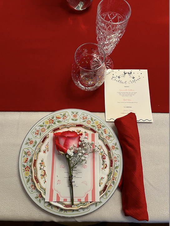 Table setting with a floral-patterned plate, gold trim, a pink flower and baby's breath on top, a red cloth napkin, a menu card, and two crystal glasses on a table with a red and cream tablecloth.
