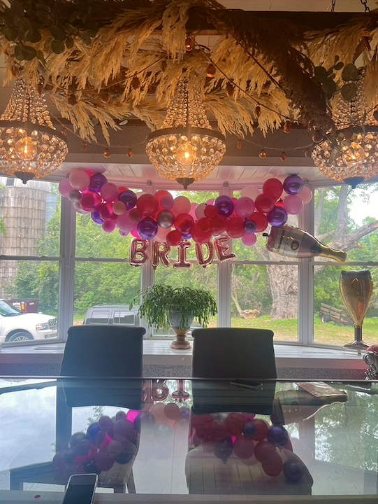 Bride celebration decoration with pink and purple balloons, balloons spelling 'BRIDE', hanging champagne bottle and glass, chandelier lighting, and a window view of trees outside.