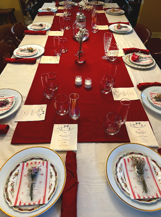 A long dining table decorated for a holiday meal with a red and white color scheme, featuring plates with holiday-themed napkins and lavender sprigs, red napkins, clear glasses, and a centerpiece with white flowers and candles.