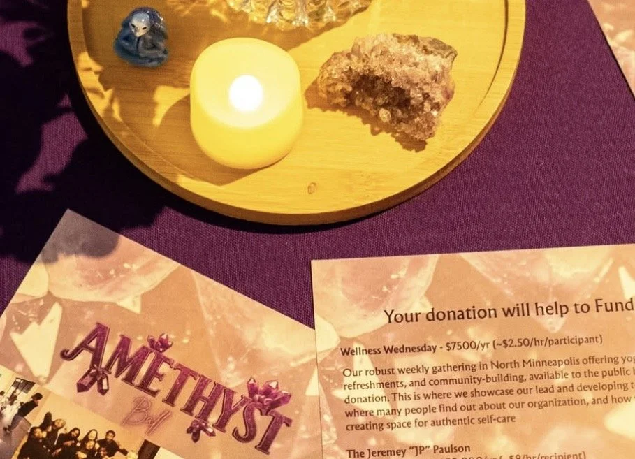 A wooden tray with a yellow LED candle, a small figurine of an angel, and a mineral or crystal specimen, placed on a purple tablecloth. There are also flyers about a fundraising event and donation information nearby.