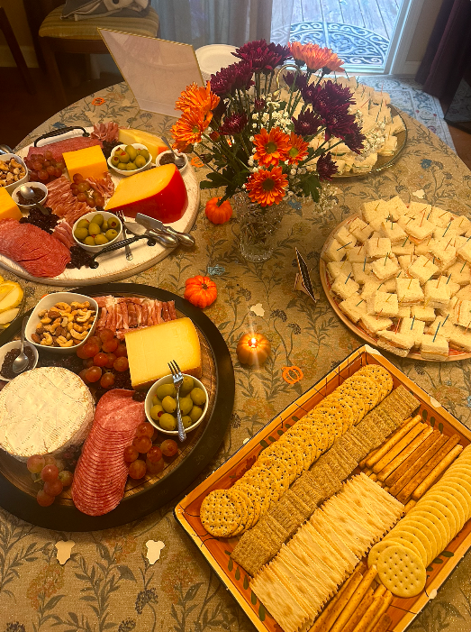 A picnic-style food arrangement on a table with cheese, meats, nuts, crackers, grapes, olives, and a floral centerpiece.