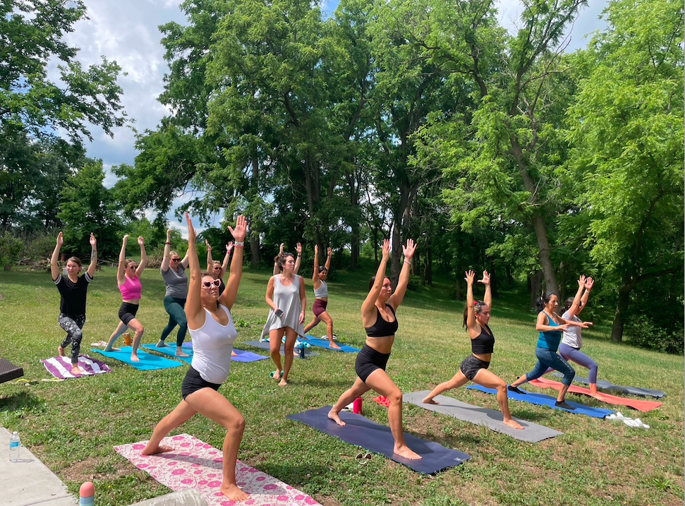 A group of women participating in an outdoor yoga class on mats in a grassy park with tall green trees in the background.