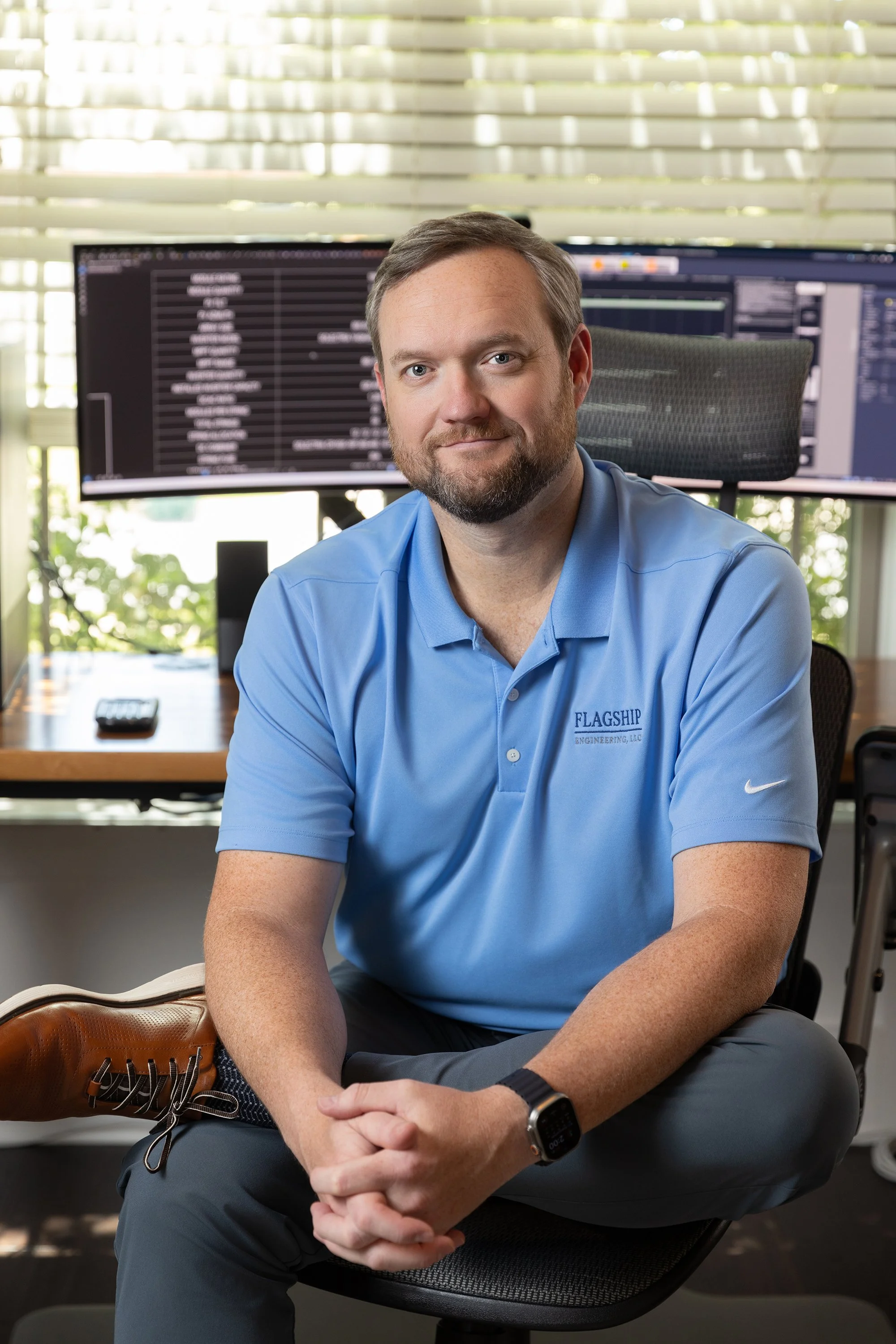 A man with a beard and short hair, wearing a blue polo shirt with an embroidered logo, sitting in an office chair with his hands clasped, in front of a desk with a computer and multiple monitors, with blinds and sunlight in the background.