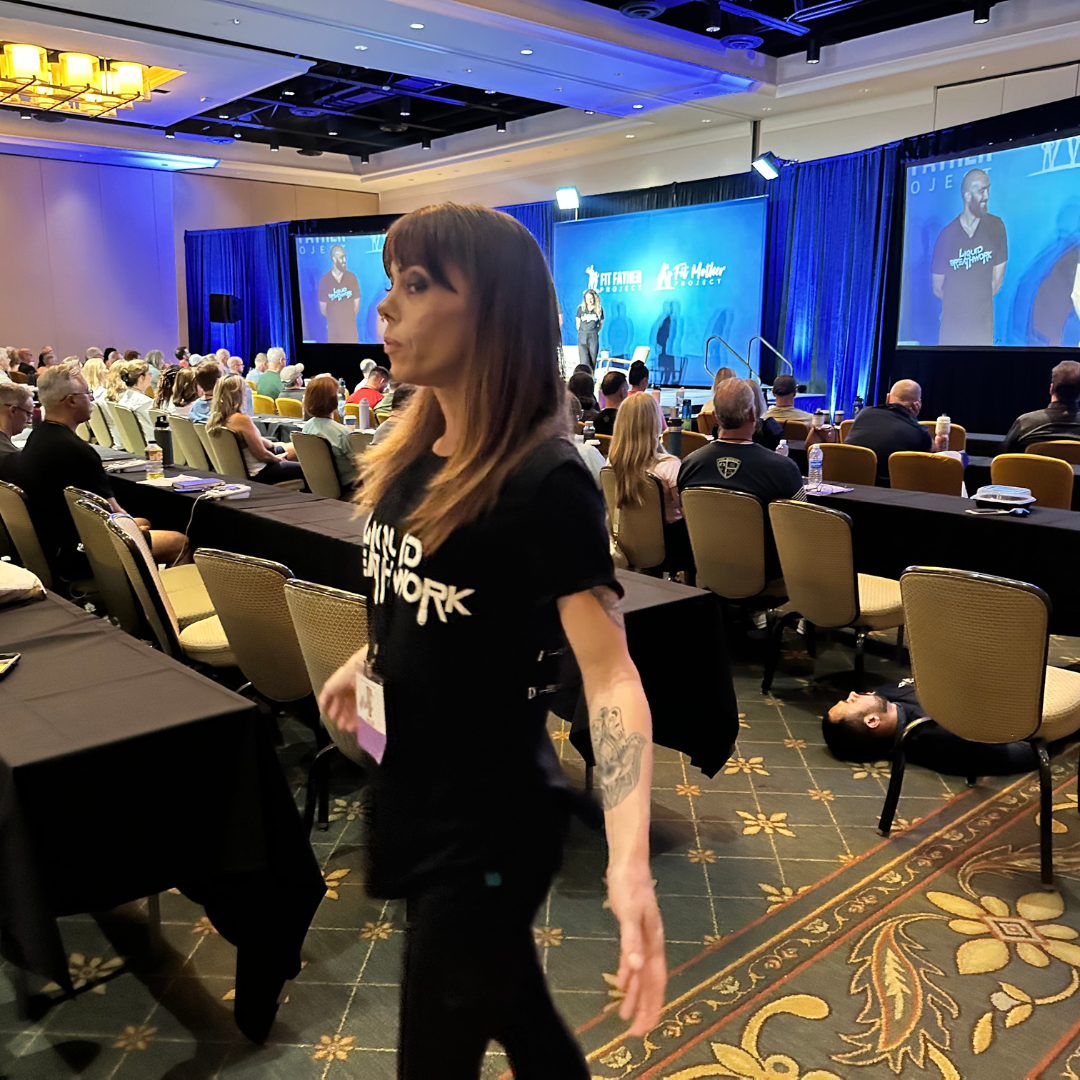 A woman with long brown hair, tattoos on her arm, walking past an audience seated at tables in a conference room, with a large stage in the background displaying a speaker and presentation slides.