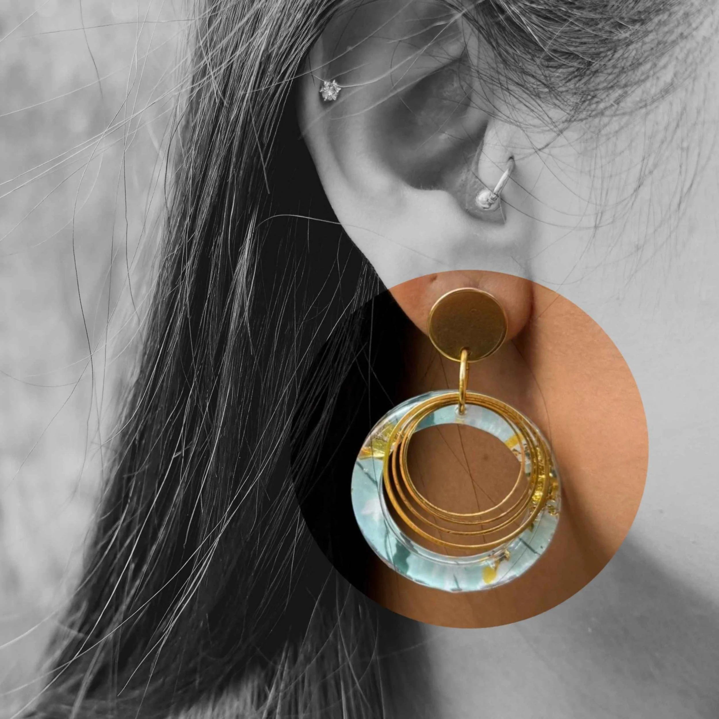 Close-up of a customer wearing circular gold and jade coloured earrings, overlaid with a design, highlighting a large statement earring with concentric gold rings - soft everyday statement style.