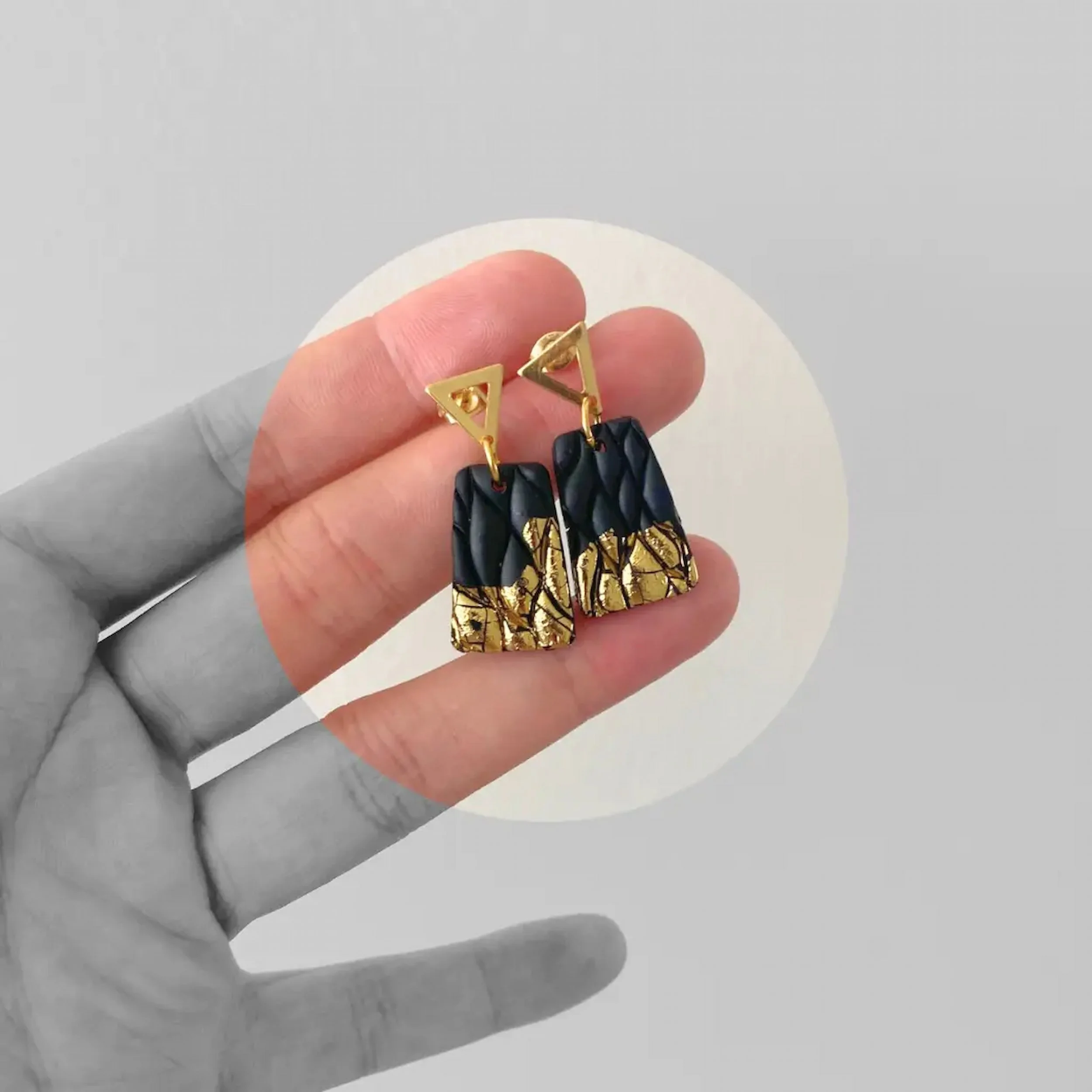 Close up of customers hand holding a pair of black and gold earrings with triangular gold studs and textured black and gold rectangular pendant - Bold wearable art piece.