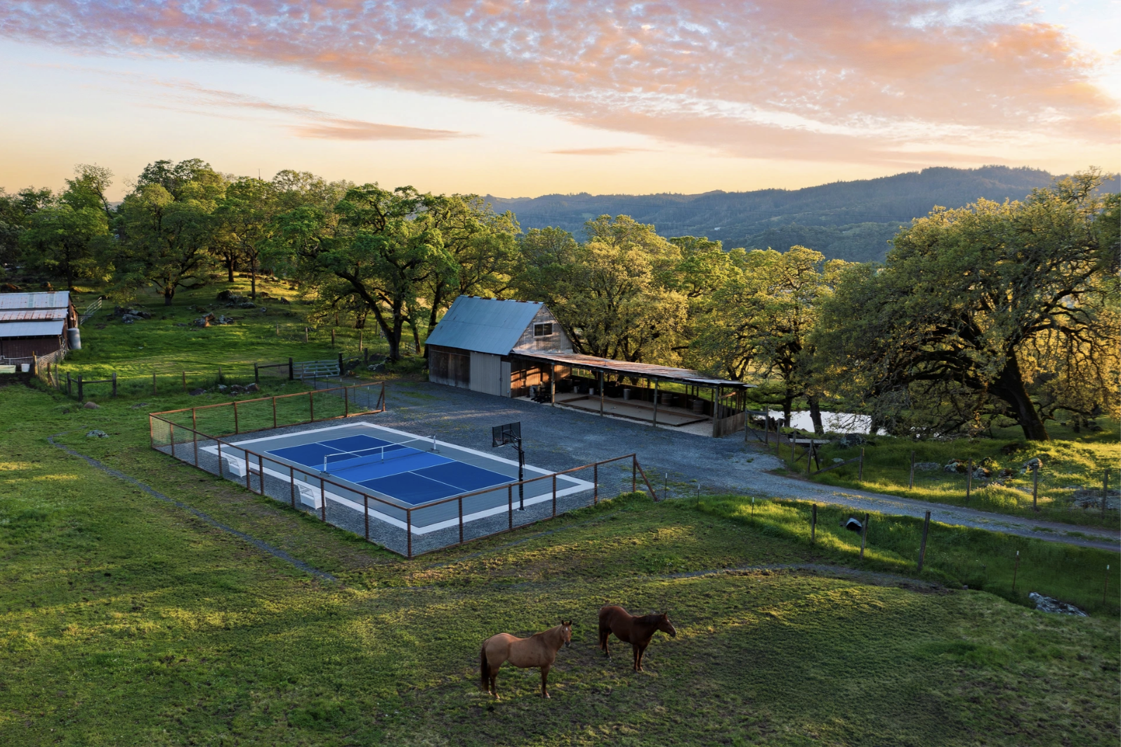 View of a countryside farm with a small tennis court, a barn, grazing horses, lush green grass, trees, and distant mountains at sunset.