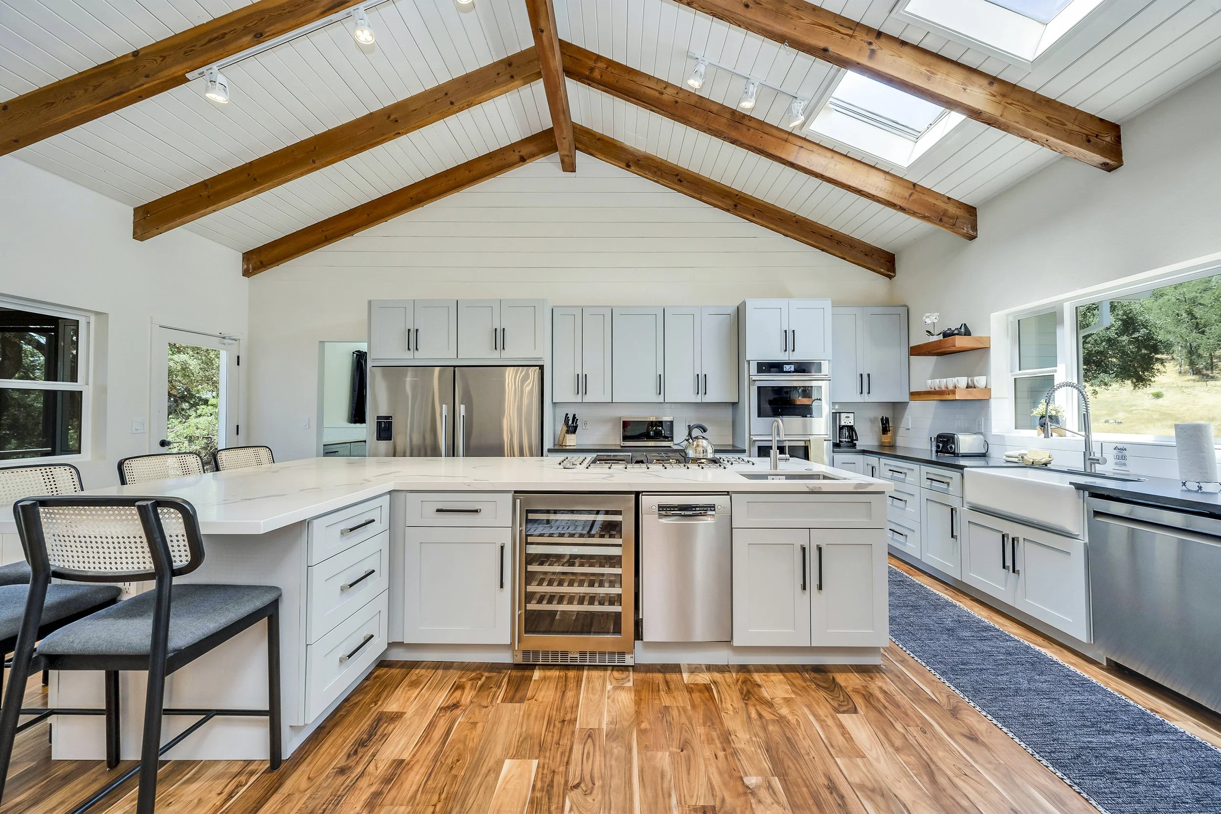 Modern kitchen with white cabinets, stainless steel appliances, wooden ceiling beams, hardwood floor, large window, and skylights.