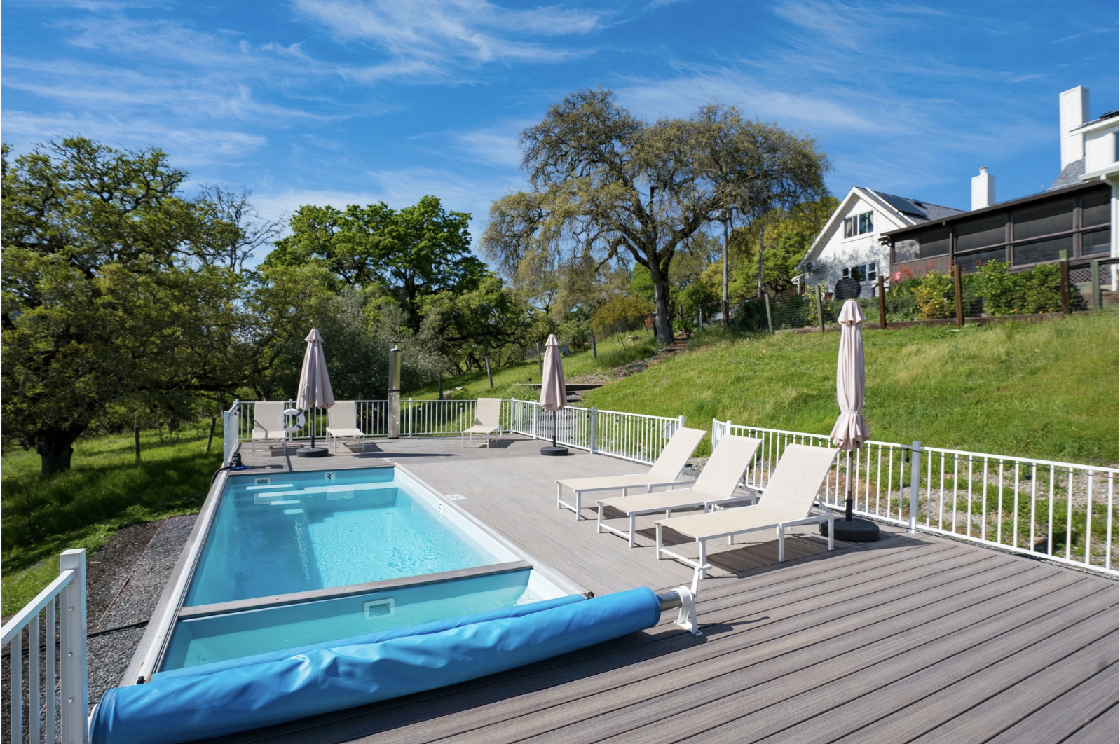 A backyard pool area with several lounge chairs, umbrellas, and a fenced yard with trees and houses on a sunny day.