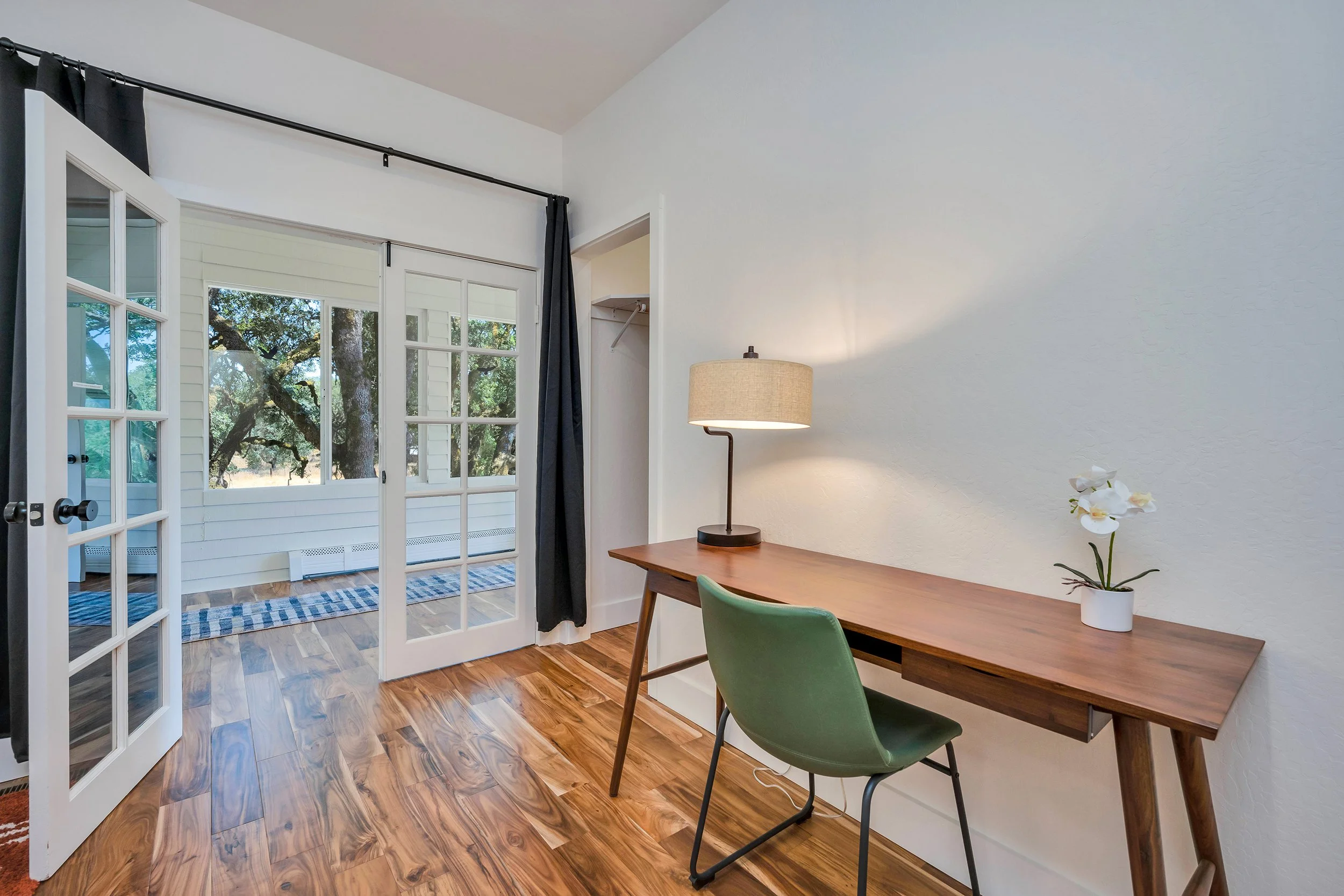 A home office with a wooden desk, a green chair, a table lamp, and a potted orchid plant on the desk. French doors lead to a porch with trees outside.