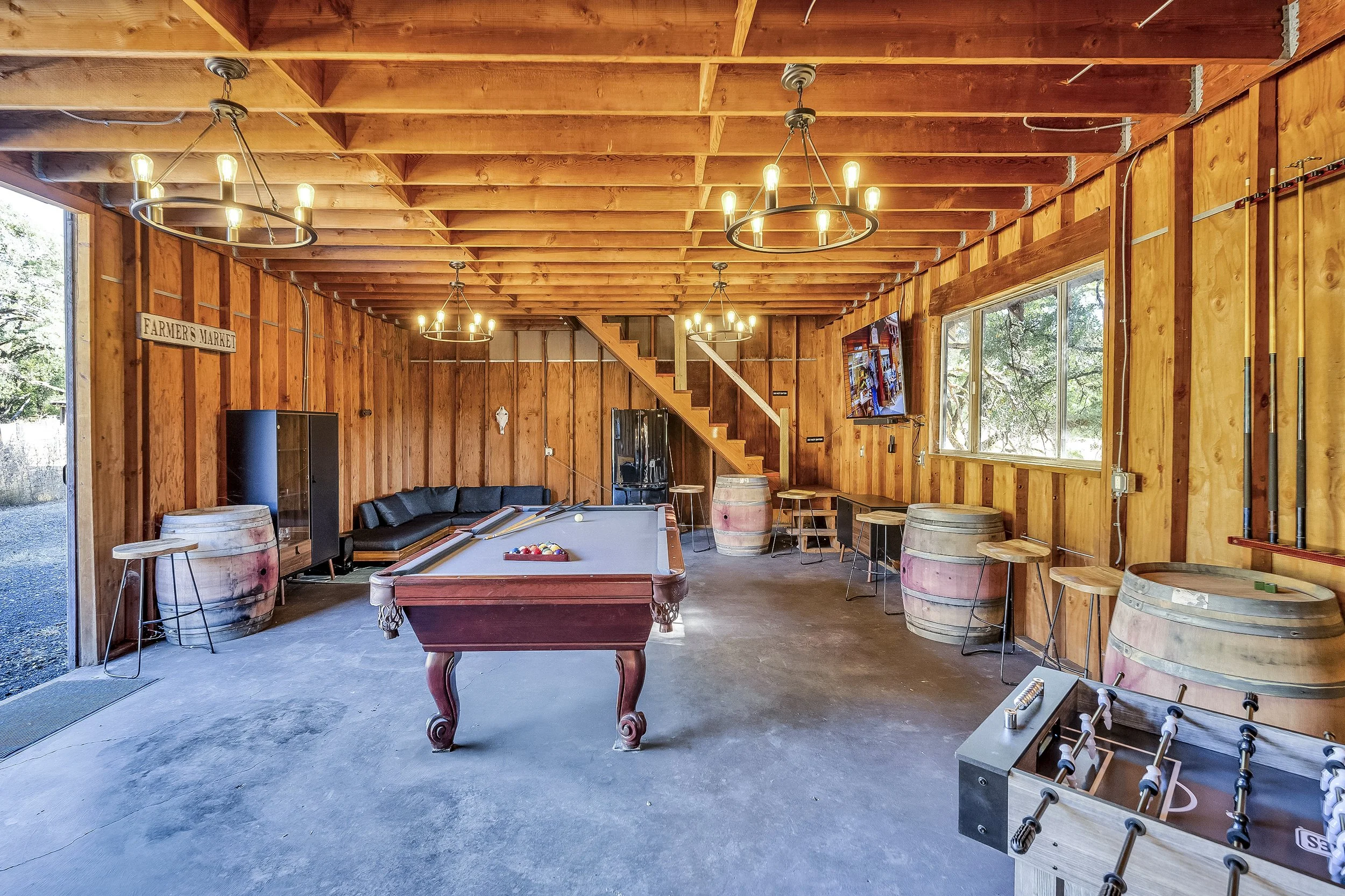 A rustic game room with wooden walls and ceiling, featuring a pool table at the center, a foosball table in the foreground, and a TV mounted on the wall. There are several barrels used as tables, bar stools, a black sectional sofa, and a staircase leading to an upper level. The room is lit by multiple chandeliers.