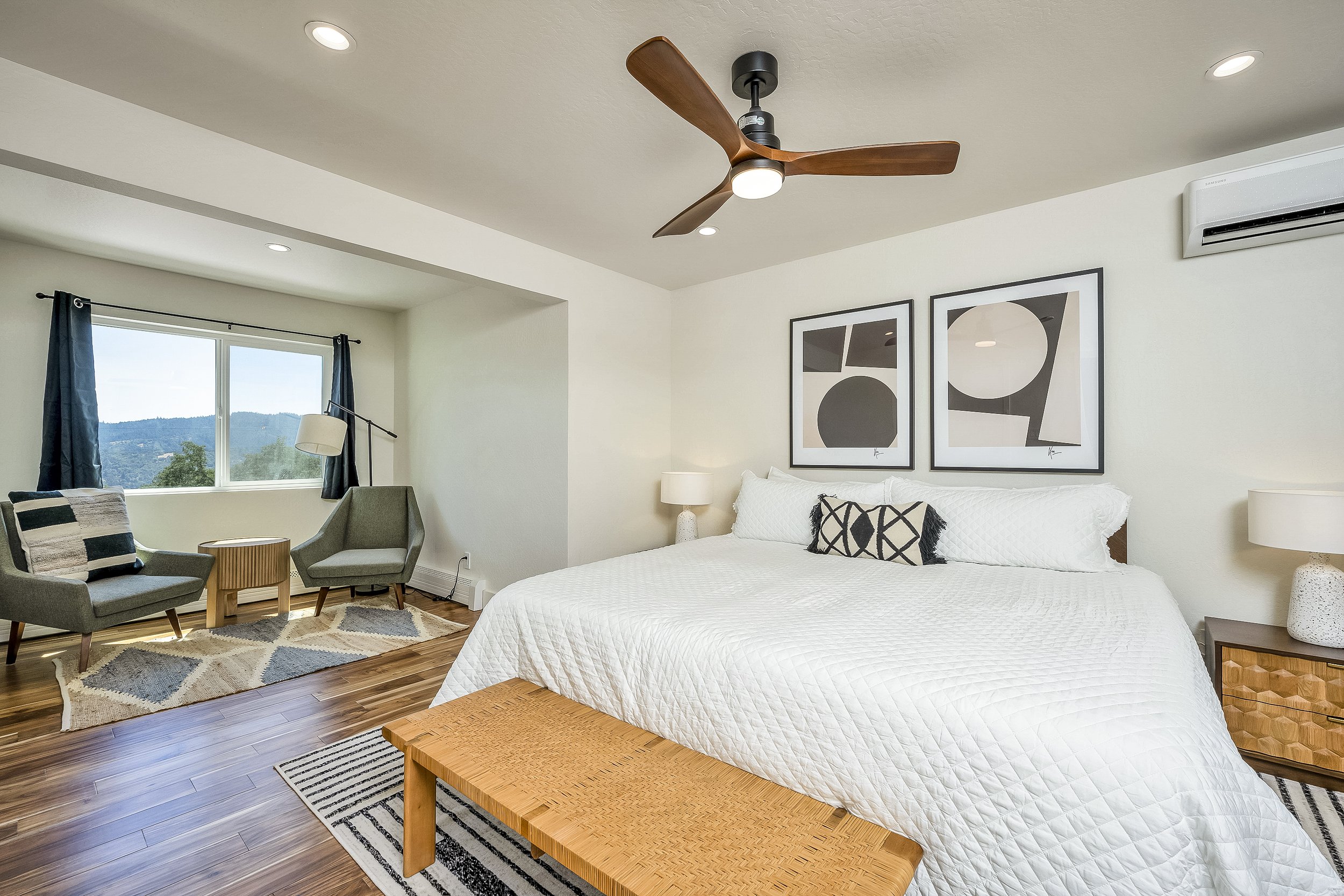Bedroom with a large bed, two framed abstract artworks above it, and a nightstand with a lamp. There is a sitting area by the window with two chairs, a small table, a lamp, and a window with blue curtains, overlooking a landscape with hills and trees.
