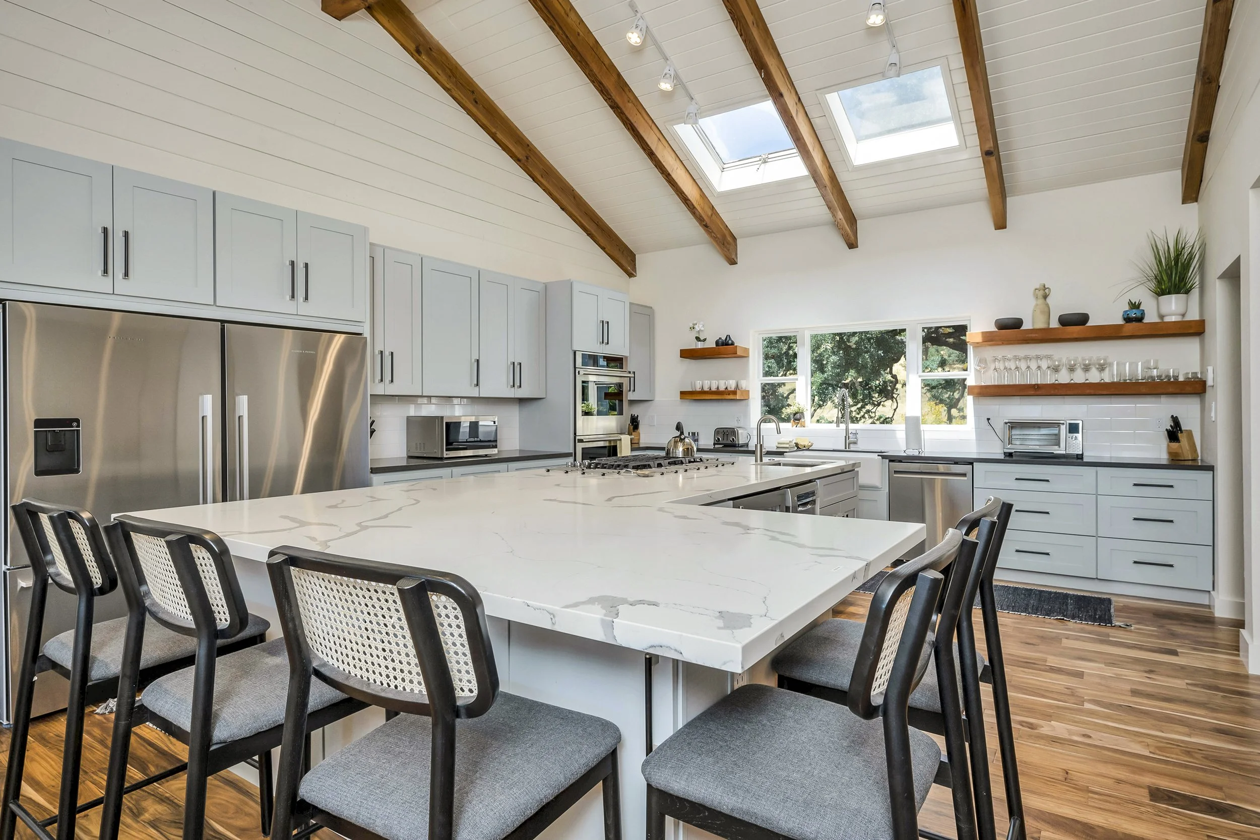 Modern kitchen with a large island countertop, gray cabinets, stainless steel appliances, open shelving with glassware, and a skylight in a wooden-beamed ceiling.