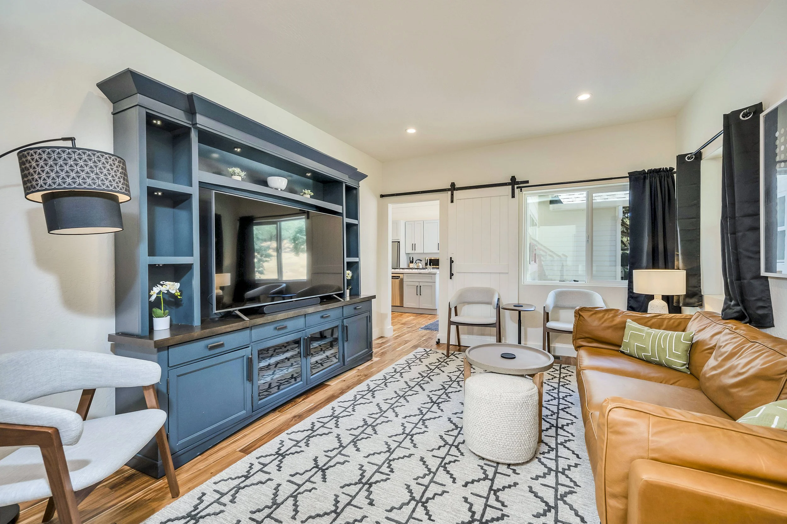 Living room with a blue entertainment center, a flat-screen TV, a leather sofa, white armchairs, and a patterned area rug. There's a doorway to the kitchen, and windows with black curtains, with hardwood floors.