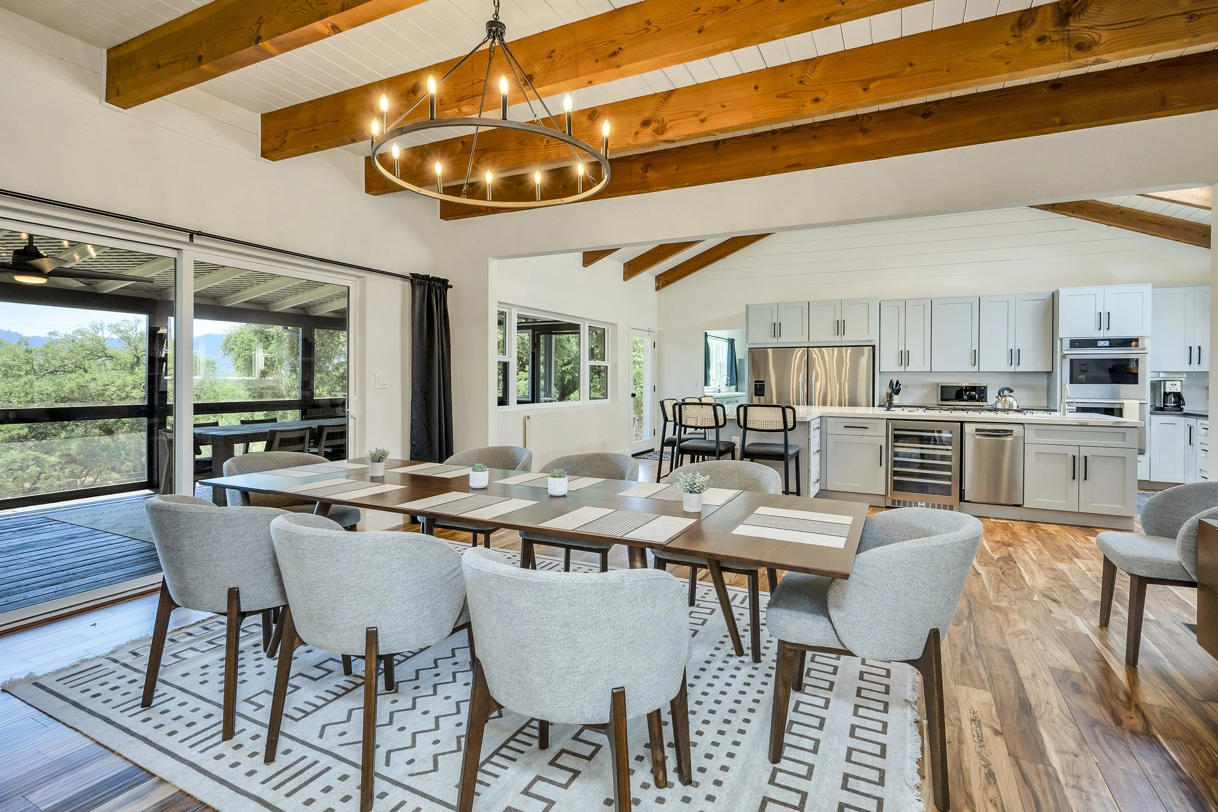 Open-concept dining and kitchen area with vaulted ceiling, wooden beams, and large windows. A wooden dining table with eight light gray chairs, a modern chandelier, and a patterned rug. Kitchen features white cabinets, stainless steel appliances, and a breakfast bar with three barstools.