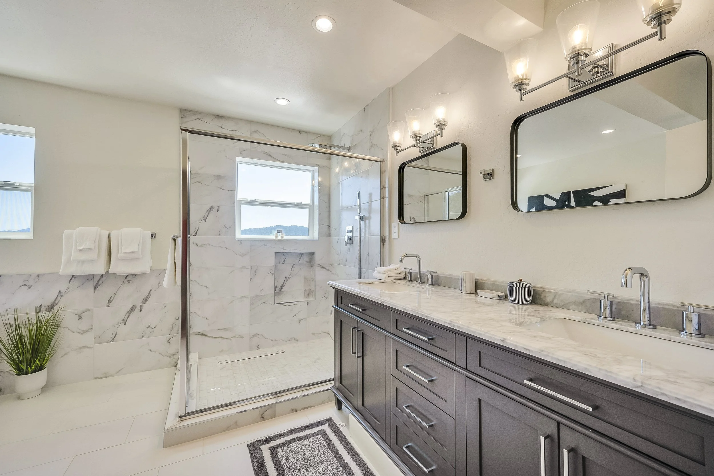 Modern bathroom with a marble-topped vanity, two black framed mirrors, elegant light fixture, walk-in shower with marble walls, and a small window overlooking a mountain landscape.