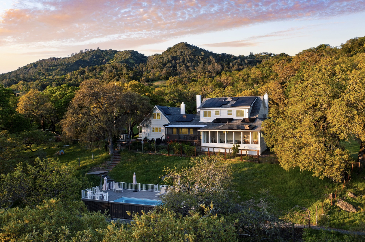 A house with solar panels on the roof, surrounded by trees, with a swimming pool and umbrellas in the backyard, set against a backdrop of green hills and a colorful sky.