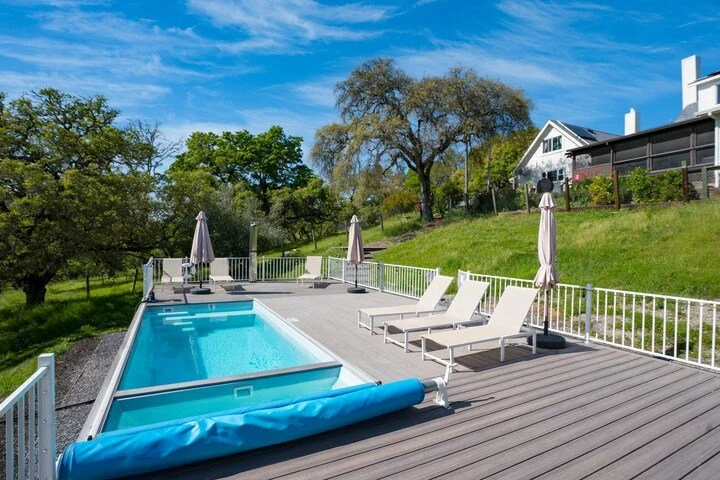 A backyard pool area with a hot tub, lounge chairs, and umbrellas on a wooden deck, surrounded by greenery and trees, under a bright blue sky.