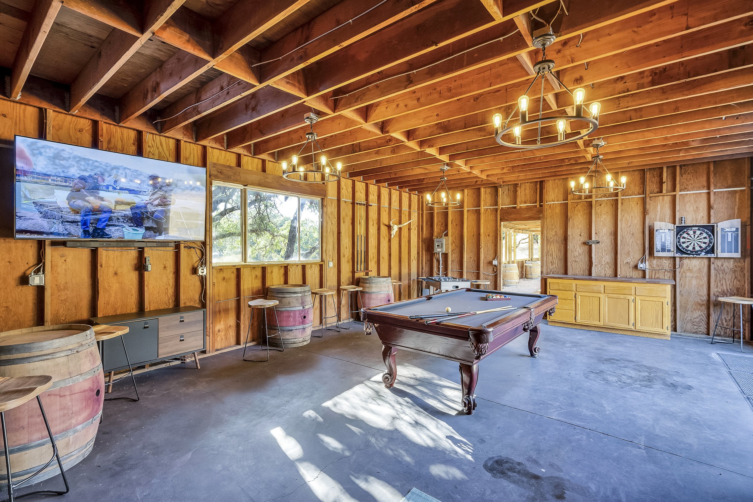 Wood-paneled game room with pool table, dartboard, flat-screen TV, and barrel tables, with chandeliers hanging from the ceiling and natural light coming through a window.