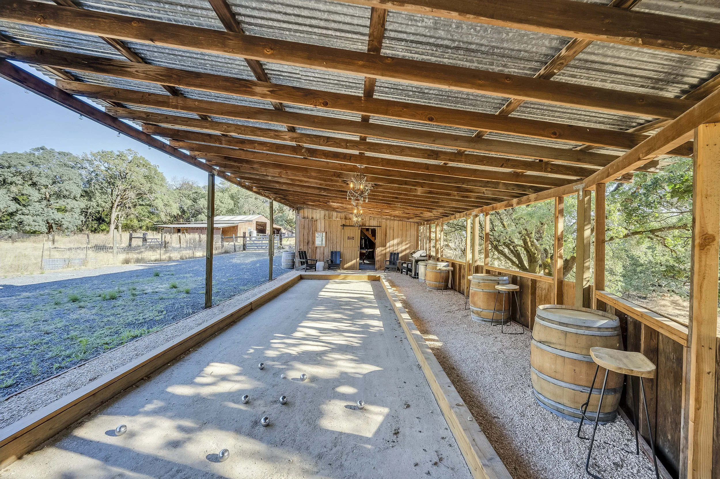 Covered outdoor bocce ball court with wooden barrels used as tables along the side, and a rustic barn in the background, surrounded by trees and open sky.