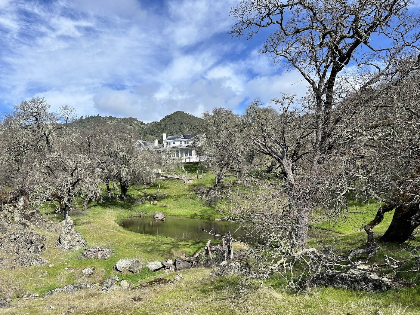 A peaceful rural landscape featuring a small pond surrounded by leafless trees, with a large white house on a hillside in the background under a partly cloudy sky.
